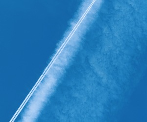 A tiny aircraft drawing two white contrails beside a vast dissolving cloud trail in deep blue sky