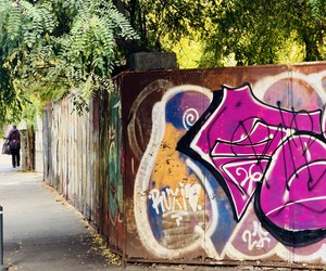 Magenta graffiti on a rusty metal fence beneath leafy trees beside a city sidewalk