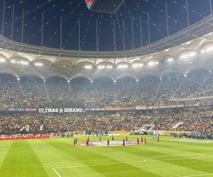 Football stadium at night with a green pitch, crowded stands, and a suspended scoreboard